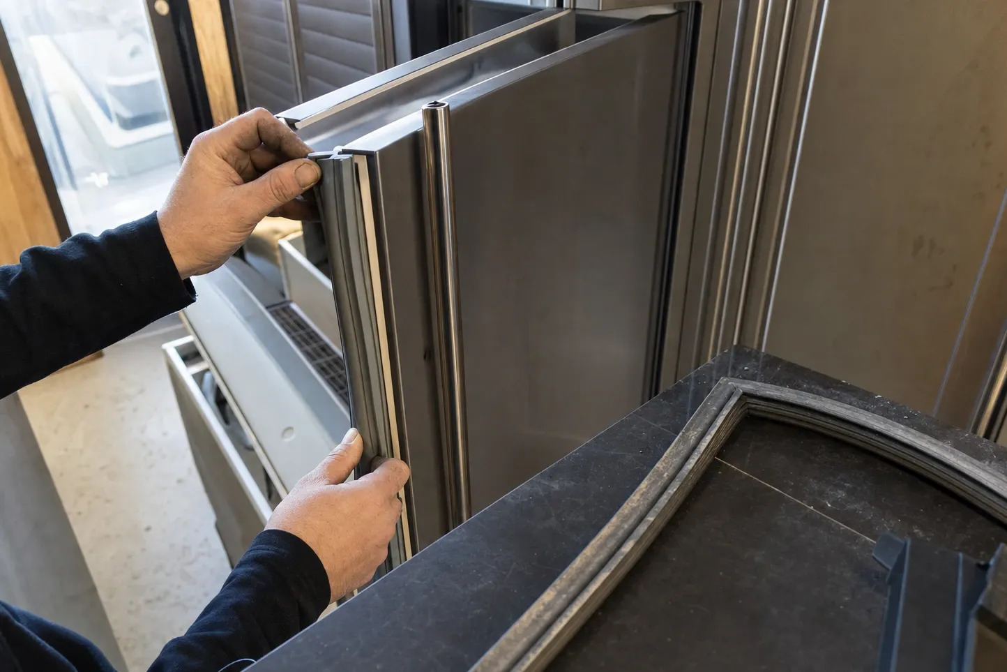 Hands easing the magnetic gasket out of the channel of a stainless door, the removed gasket resting on the counter