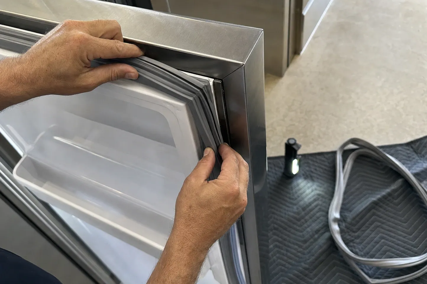 Hands pressing a new magnetic gasket into the channel of a stainless door, the old gasket coiled on the mat below